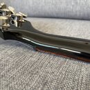 Detailní pohled na hřbet černého lakovaného krku kytary a hlavu s kovovými ladičkami. Lakovaný povrch je hladký a vysoce lesklý, v dolní části je vid…