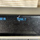 Detail černého efektového pedálu s texturou povrchu. Na horní hraně jsou modrým písmem nápisy GUITAR IN a PEDAL 2. Nad nápisem PEDAL 2 je viditelné o…