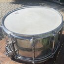 Stříbrný kovový buben s bílou blánou. Na bláně je vidět černé logo, které je však nečitelné. Povrch kovového korpusu vykazuje známky opotřebení v pod…