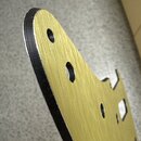 Detailní záběr na zlatý kovový plech, pravděpodobně pickguard k elektrické kytáře. Povrch má jemnou broušenou texturu a v něm jsou vyvrtány otvory. O…