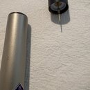 Stříbrný kovový válcový předmět s modrobílým logo NEUMANN. Vedle něj je položen černý plastový kryt s vyčnívajícím kovovým hrotem. Povrch stříbrného…