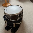 Tmavě hnědý snare drum s kovovým obručováním, umístěný na černém transportním pouzdře. Na bílém bláně jsou viditelné nápisy REMO a YAMAHA. Nástroj vy…