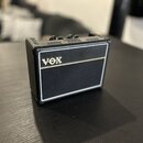 Malý kytarový zesilovač s černým povrchem z texturovaného materiálu a bílými lemovými pruhy. Na přední straně je umístěno zlaté logo VOX. Horní panel…