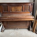 Hnědé dřevěné piano s výraznou masivní konstrukcí a dekorativními panely. Povrch je pokryt prachem a vykazuje známky opotřebení v podobě škrábanců. N…