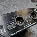 Černý kovový přístroj s broušeným povrchem a dvěma XLR výjemy. Na šasi jsou nápisy OUTPUT 1 a OUTPUT 2, u každého z nich se nachází přepínač s označe…