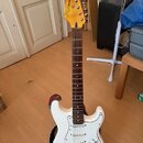 Bílá elektrická kytara s tmavým hnedým hmatníkem a bílým pickguardem. Na hlavě je žlutý lak a nápis "Wayland HEADLESS Standard". Kytara je umístěna v…
