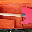 Růžová elektrická kytara v otevřeném kufru s oranžovou výstelkou. Na zadní straně krku je viditelné logo Fender Custom Shop. Kytara má světle dřevěný…