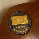 Detail zadní strany kytary s hnědým dřevěným povrchem. Větší černý kruhový kovový prvek obsahuje zlatou plošku s nápisem Gibson CUSTOM a texty EST. 1…
