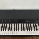Černý elektronický klavír s bílými a černými klávesami. Na ovládacím panelu jsou posuvníky, tlačítka a nápisy včetně VOLUME, LOW, MIDDLE, HIGH, VOICE…