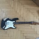 Elektrická kytara s černým lakem, který vykazuje výrazné opotřebení a odrolení v horní části těla. Nástroj má bílý pickguard, hnědý hmatník a světle…