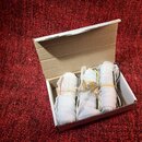 V otevřené bílé kartonové krabičce jsou tři předměty zabalené v bílém pěnovém materiálu a zajistěné žlutými gumičkami. Předměty mají válcový tvar a j…