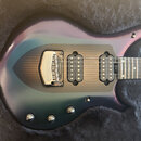 Elektrická kytara s iridescentním povrchem v odstínech fialové a zelené, položená v černém plišovém kufru. Nástroj má dvě humbuckery, karbonový pickg…