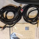 Černý pletený kabel s kovovými konektory a červeným prstencem u jednoho z nich. Na černém plastovém doplňku je nápis SENNHEISER. Vedle kabelu leží pa…
