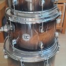 Tři bubny s hnědým mramorovaným povrchem naskládané na sebe. Každý buben má chromované obruče a mechanismy, přičemž všichni nesou logo DC DRUMS. Na h…