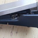 Detailní pohled na část černého plastového pedálu s kovovou stříbrnou plošinkou. Na povrchu nejsou viditelné žádné značky ani nápisy. Předmět leží na…