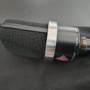 Kondenzátorový mikrofon s černým mřížkovým hlavcem a stříbrným kovovým prstencem. Na černém těle je vidět červené trojúhelníkové logo Neumann a bílé…