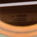 Detail vnitřku akustického nástroje s viditelnou dřevěnou štítkovou nálepkou. Na štítku je černé logo a nápis YAMAHA, pod kterým jsou texty MADE IN T…