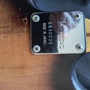 Detail zadní strany kytary s kovovým krytem v oblasti spojení krku s tělem. Na krytu je vyryto logo Ibanez, kód E830030 a nápis MADE IN JAPAN. Povrch…