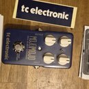 Modrý kytarový efekt s nápisy tc electronic, MOJOMOJO a PAUL GILBERT OVERDRIVE. Pedal má čtyři bílé ovládací knoflíky označené jako BASS, TREBLE, DRI…