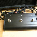 Černý nožní spínací pedalboard s třemi kovovými spínači. Nad spínači jsou čísla 1, 2 a 3 s doprovodnými kontrolkami v červené, žluté a modré barvě. V…