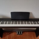 Černý digitální piano v černém stojanu s třemi pedály. Na centerXu ovládacího panelu a na hudebním stojanu je viditelné logo YAMAHA. Nástroj vykazuje…