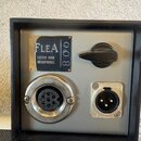 Štihlé šedé zařízení v černém rámu s dvěma konektory. Na horní části je kovový štítek s nápisem FLEA CUSTOM MADE MICROPHONES a číslem 008. Vpravo je…