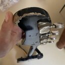 Detail hlavy černé strunové nástroje s kovovými ladičkami. Horní hrana hlavy vykazuje výrazné opotřebení a odloupení laku, pod kterým je vidět světlé…