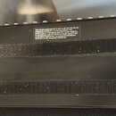 Fotografie zachycuje spodní stranu černého elektronického zařízení. Na povrchu jsou dva dlouhé, vodorovné prouhy lepicího suchého zapojení. V horní č…