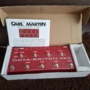 Červený efektový spínací pedalboard v kartónové krabici s polystyrenovou výplní. Na zařízení je bílý nápis OCTA-SWITCH MK3 a v dolní části text MADE…