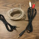 Na dřevěném povrchu leží tři audio kabely. Jeden je bílý s odrezvěnými konci vodičů, druhý je černý s dvěma 3,5mm jack konektory a třetí je černý s j…