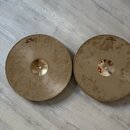 Dvě kovové činely v zlatavé barvě položené na světlém laminátovém povrchu. Levý činele má nápis 12"/30cm HIHAT-TOP a pravý činele má nápis 15"/38cm H…