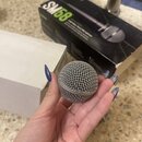 Ruka drží kovovou mřížku mikrofonu v šedé barvě. V pozadí je vidět černý karton s nápisy SM58 a Legendary Vocal Microphone. Vedle nich je bílá krabic…