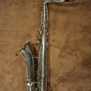 Stříbrný saxofon s černým mouthpiecem. Povrch nástroje vykazuje viditelné opotřebení, škrábance a oxidaci. Na těle nástroje nejsou čitelné žádné znač…