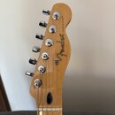 Fotografie zachycuje hlavu kytary z přírodního světlého dřeva s chromovanými ladičkami. Na povrchu je vidět bílý nápis Fender a TELECASTER®. V dolní…