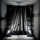 Fotografie zobrazuje studio pro focení produktů s černým pozadím a šedou podlahou. V prostoru jsou umístěny dvě černé kovové police s integrovaným os…