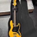 Žlutá basová kytara v černém měkkém pouzdře. Na hlavě nástroje je viditelné logo "Squier". Kytara má černý pickguard a čtyři struny. Povrch nástroje…
