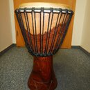 Djembe s tělem z tmavého dřeva a napnutou světlou kůží. Nástroj je natažen černým provázkem, který tvoří mřížkovou strukturu. Spodní část dřevěného t…