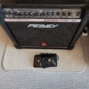 Černý kytarový kombo zesilovač s nápisy TRANSTUBE SERIES, STUDIO PRO 112 a PEAVEY. Na spodní části mřížky je červené logo ve tvaru stylizedého písmen…