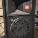 Černý reproduktor v bedně potažené látkou s plastovými rohy. V horní části je umístěn roh s oranžovým logem JBL a nápisem PROFESSIONAL PRODUCTS. Spod…