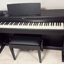 Černý digitální piano s bílými a černými klávesami. Na horní hraně je umístěno logo YAMAHA. Součástí je černá stolička a na nástroji leží černý napáj…