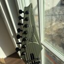 Detail hlavy elektrické kytary v olivově zelené barvě s matným povrchem. Na hlavě je bílý nápis K-7 Series a logo Ibanez. Nástroj má sedm ladicích ko…