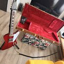 Červená elektrická kytara s bílým ráfkem a hnědým hmatníkem. Na hlavě je viditelné logo Gibson. Vedle kytary je otevřený hnědý kufr s červeným vnitřk…