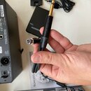 Ruka drží černý kabel s konektory XLR a 6,35mm jack. Na těle jackového konektoru je vyražen nápis NEUTRIK. V pozadí jsou vidět černý přístroj s nápis…