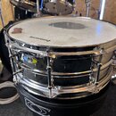 Černý snare drum s chromovaným obručováním a napínacími šrouby. Na bílém bubnovém povrchu je v horní části hnědá skvrna a v dolní části nápis "Ludwig…