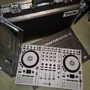Bílý DJ kontrolér s nápisem TRAKTOR KONTROL S4. Vedle něj je položen hnědý karton s nápisem TRAKTOR KONTROL S4 a v pozadí černý transportní kufr s lo…