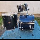 Sada černých bubnů s kovovým chassis. Sestava obsahuje basový buben, tom a malý snare umístěný na stojanu. Na tomu je vidět zlatavý oválný štítek s n…