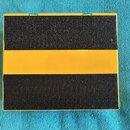 Kvadratické etui s černým, texturovaným povrchem a širokým žlutým pruhem uprostřed. Předmět je položen na modrém textilu. Na povrchu nejsou viditelné…