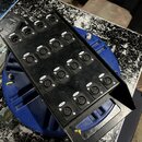 Černý kovový rozdělovač s 20 konektory XLR. Každý konektor je opatřen malým bílým štítkem s číslem od 1 do 20. Zařízení leží na modrém plastovém návi…