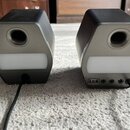 Dva identické kompaktní reproduktory v šedé a černé barvě s bílými předními panely. Na zadní straně pravého reproduktoru jsou viditelné konektory USB…
