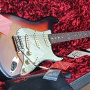 Elektrická kytara s tónem těla sunburst a bílým pickguardem v černém kufru s červenou výplní. Na hlavě hmatníku je viditelné logo Fender. Kytara má t…